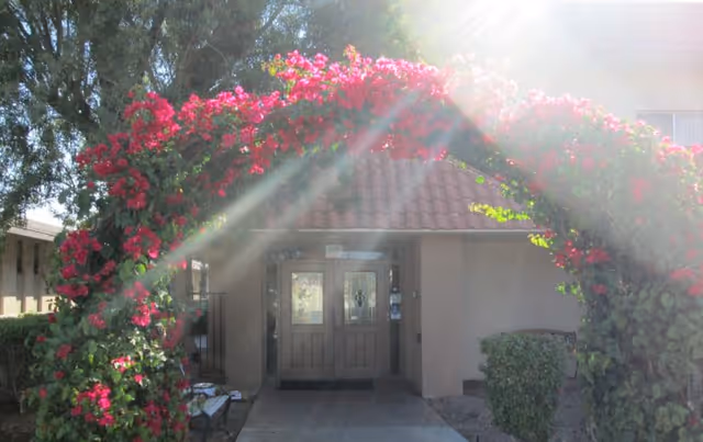 Entrance to a building with double wooden doors under a red-tiled roof, framed by an arch covered in vibrant pink flowers and greenery, with sunlight streaming through the foliage.