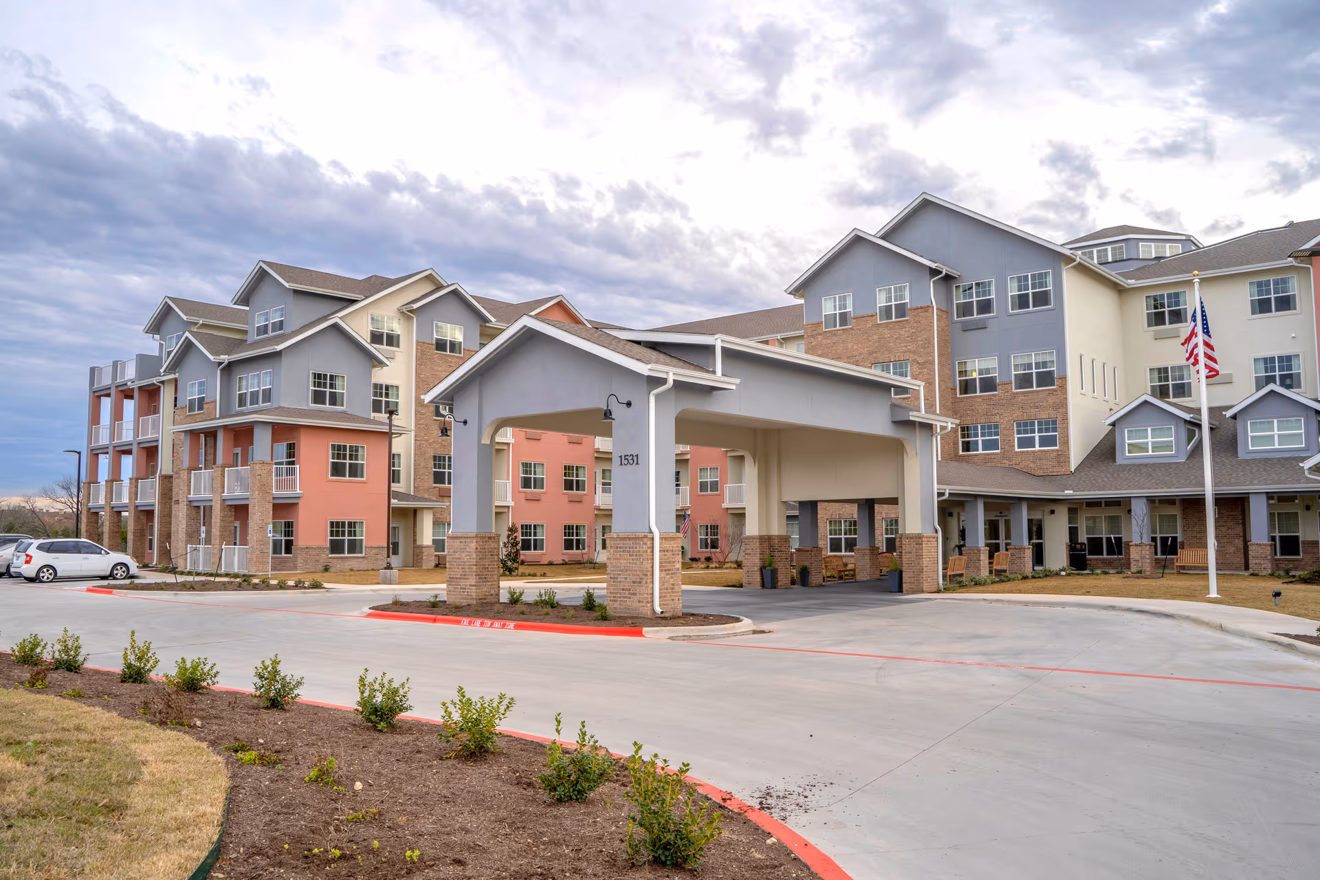 Front exterior of a multi-story senior living building with a covered entrance/porte-cochère and an American flag.