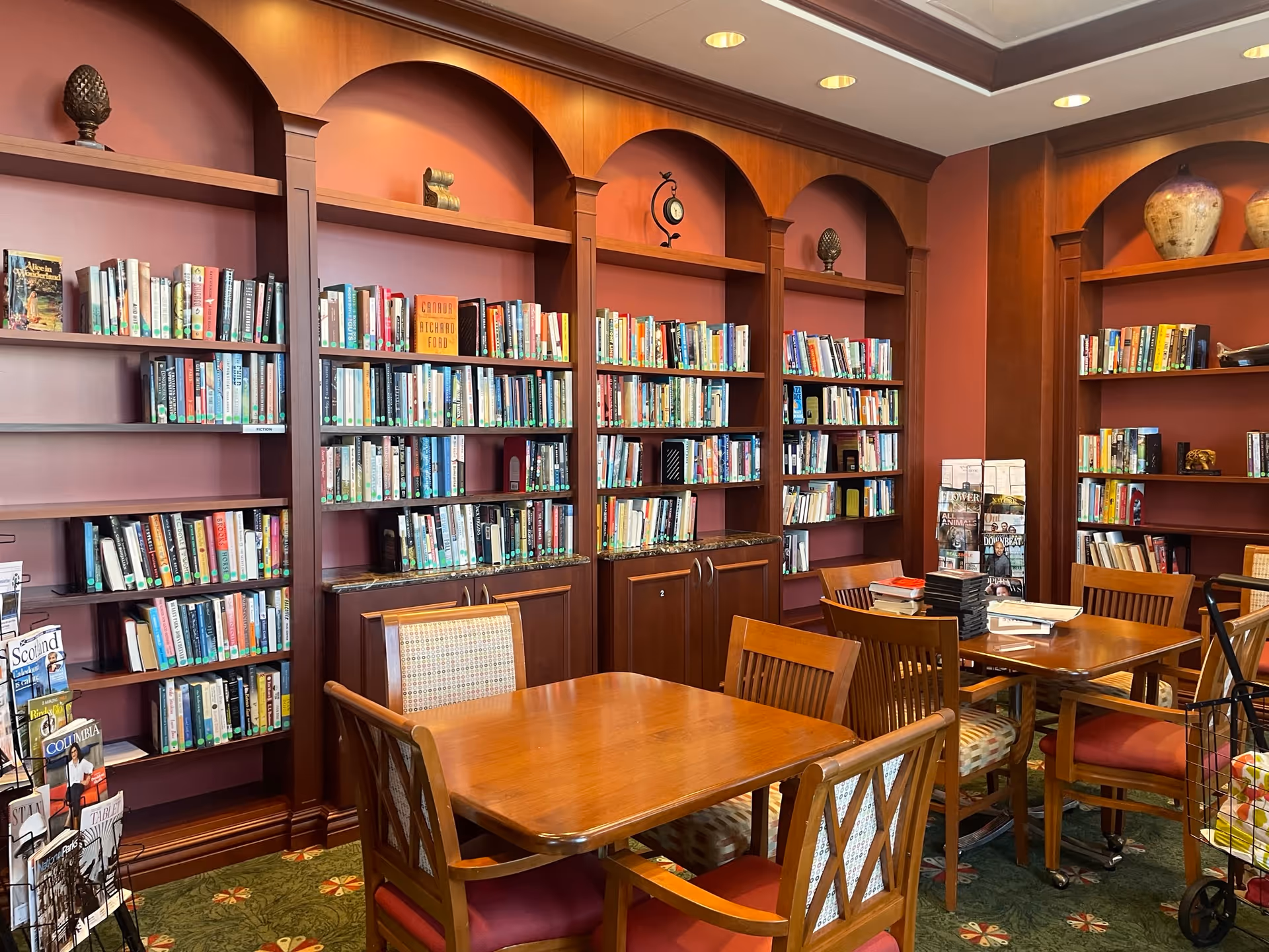 Interior of a library or reading room with wooden bookshelves filled with books against red walls. There are wooden tables and chairs arranged in the center of the room, with some books and magazines on the tables. The room has a green carpet with a floral pattern and decorative items on the top shelves.
