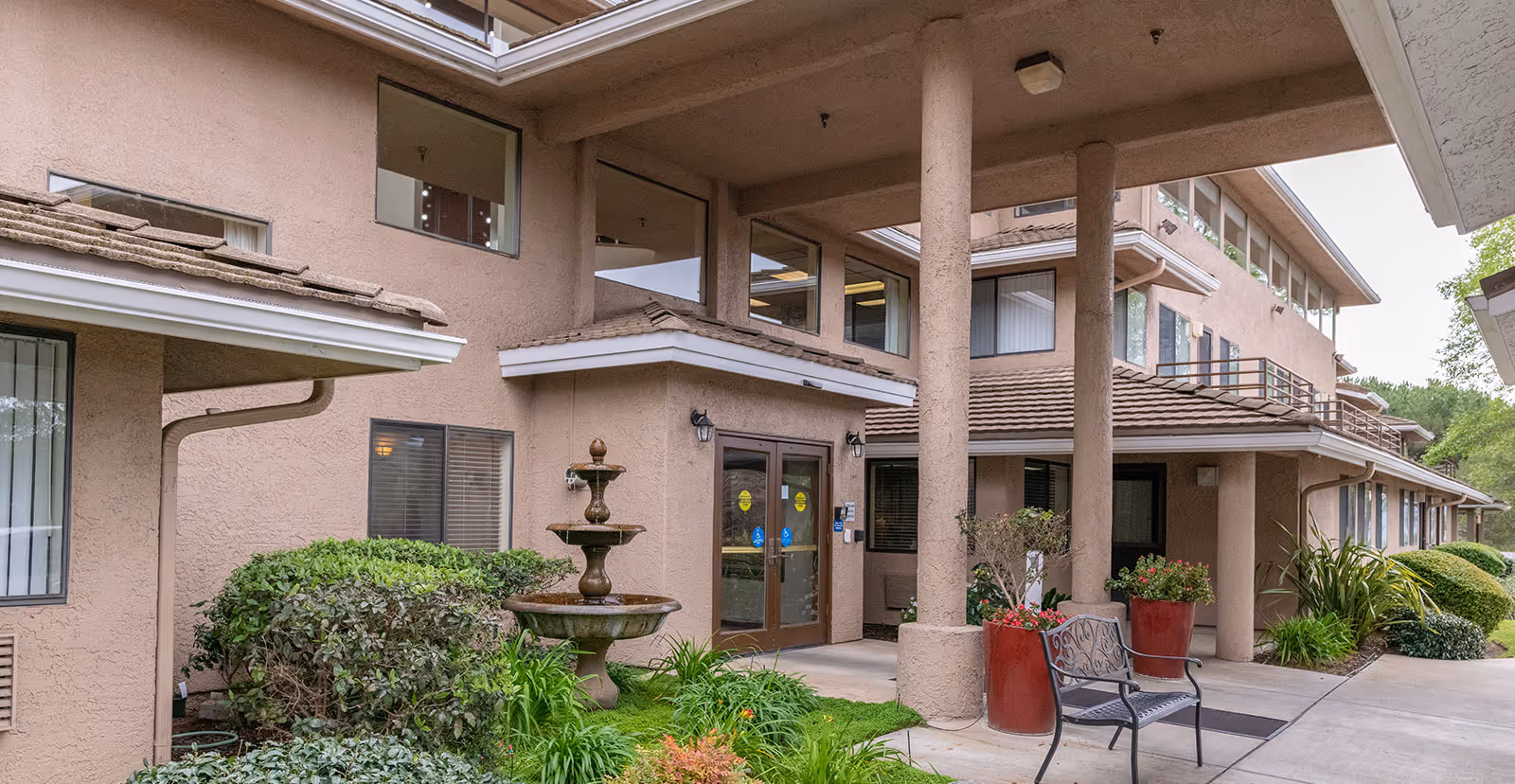 Exterior view of the entrance to Las Brisas Retirement Community, featuring a covered walkway supported by columns, a three-tiered water fountain, potted plants, bushes, and a metal bench near the doorway.