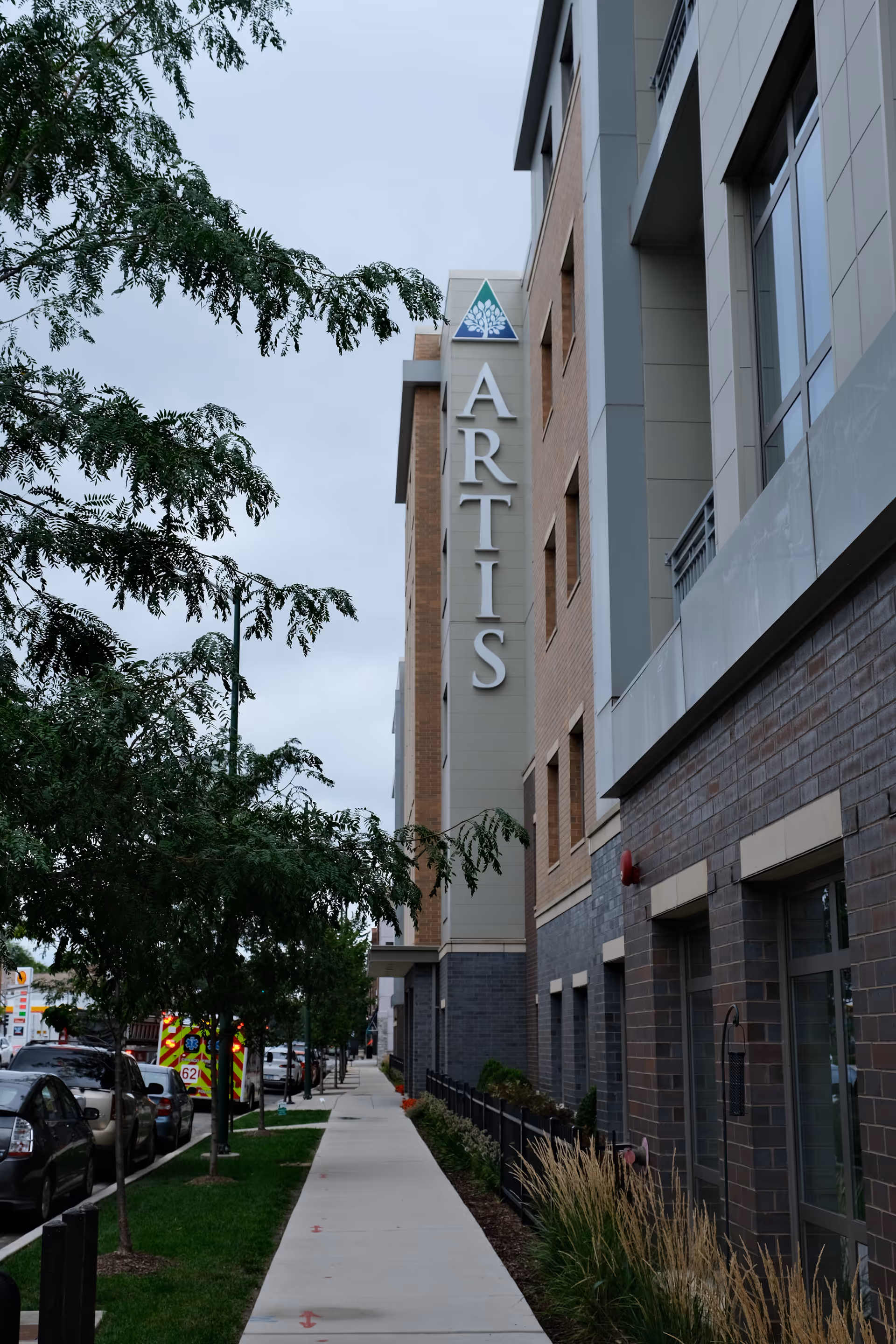 Sidewalk view along a modern multi-story building with the large vertical sign 'ARTIS' on the facade. Trees line the sidewalk and several parked cars are visible along the street. The sky is overcast.