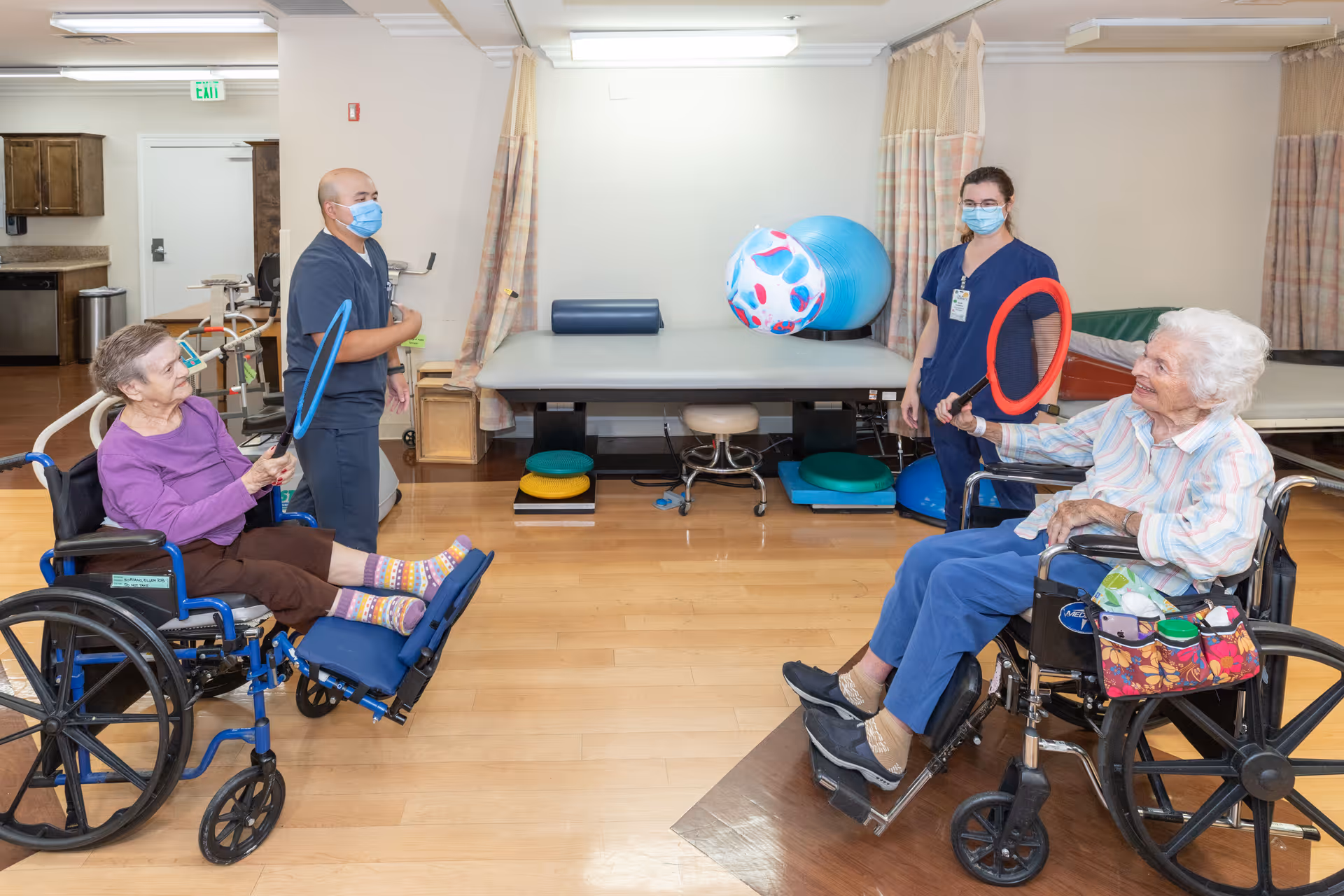 Two elderly women in wheelchairs playing a game with ring toss paddles and a large inflatable ball in a rehabilitation or therapy room, assisted by two healthcare workers wearing masks.