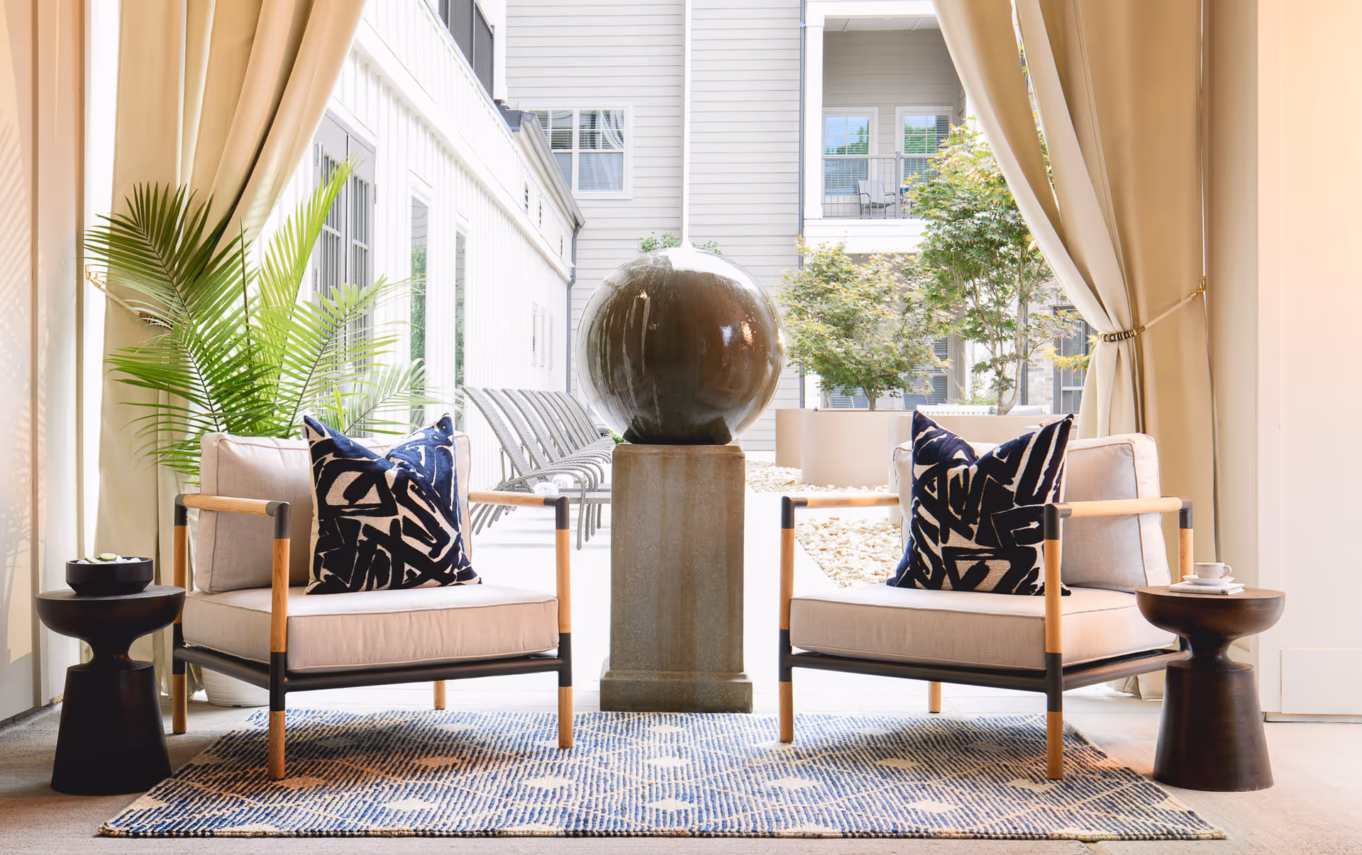 Two cushioned armchairs with patterned pillows are placed on a blue and white rug in a bright indoor seating area. Between the chairs is a pedestal with a large spherical water feature. There are small round side tables next to each chair, one with a small plant and the other with a cup and saucer. Beige curtains frame the space, and large windows and greenery are visible in the background.