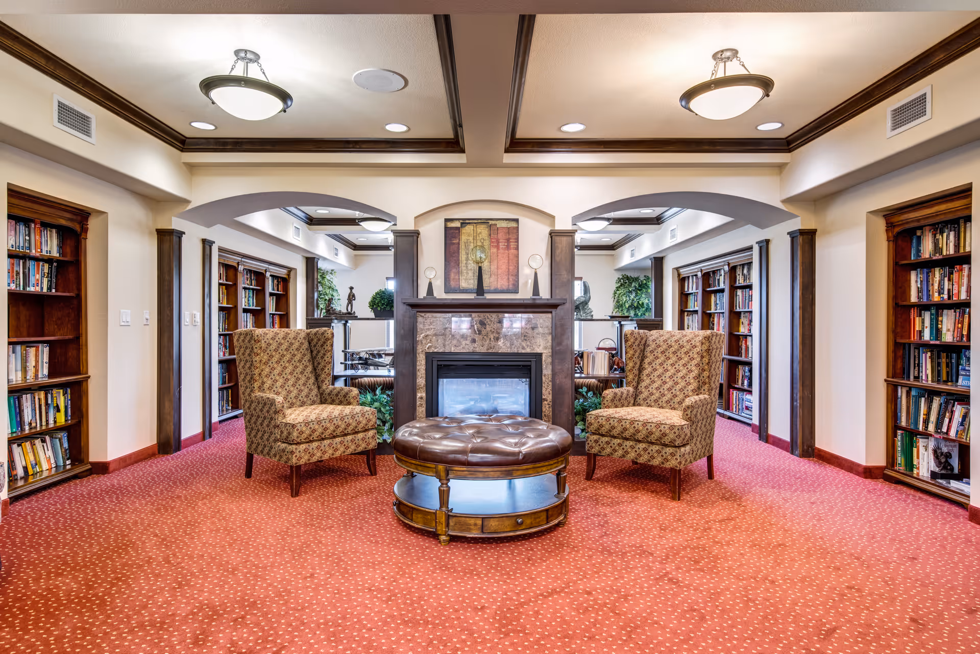 A cozy interior common area with two patterned armchairs facing a round leather ottoman in front of a fireplace. The room features built-in wooden bookshelves filled with books on both sides, red carpet flooring, and decorative ceiling lights. There are plants and artwork above the fireplace, creating a warm and inviting atmosphere.