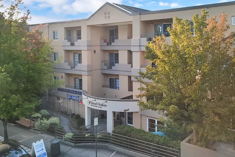 Exterior view of a three-story assisted living facility named Island House Assisted Living, with balconies on each floor, surrounded by trees and a sidewalk with benches and signage.