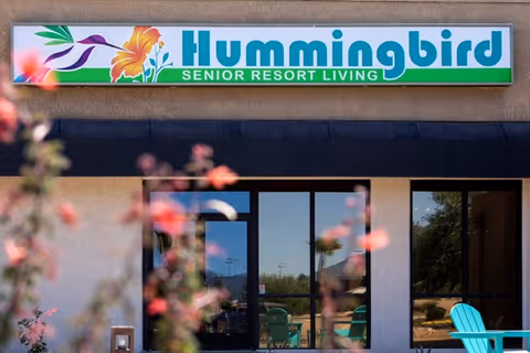 Exterior view of Hummingbird Senior Resort Living facility showing the building entrance with glass doors and windows, outdoor chairs, and a sign above the entrance with a hummingbird and flower graphic.