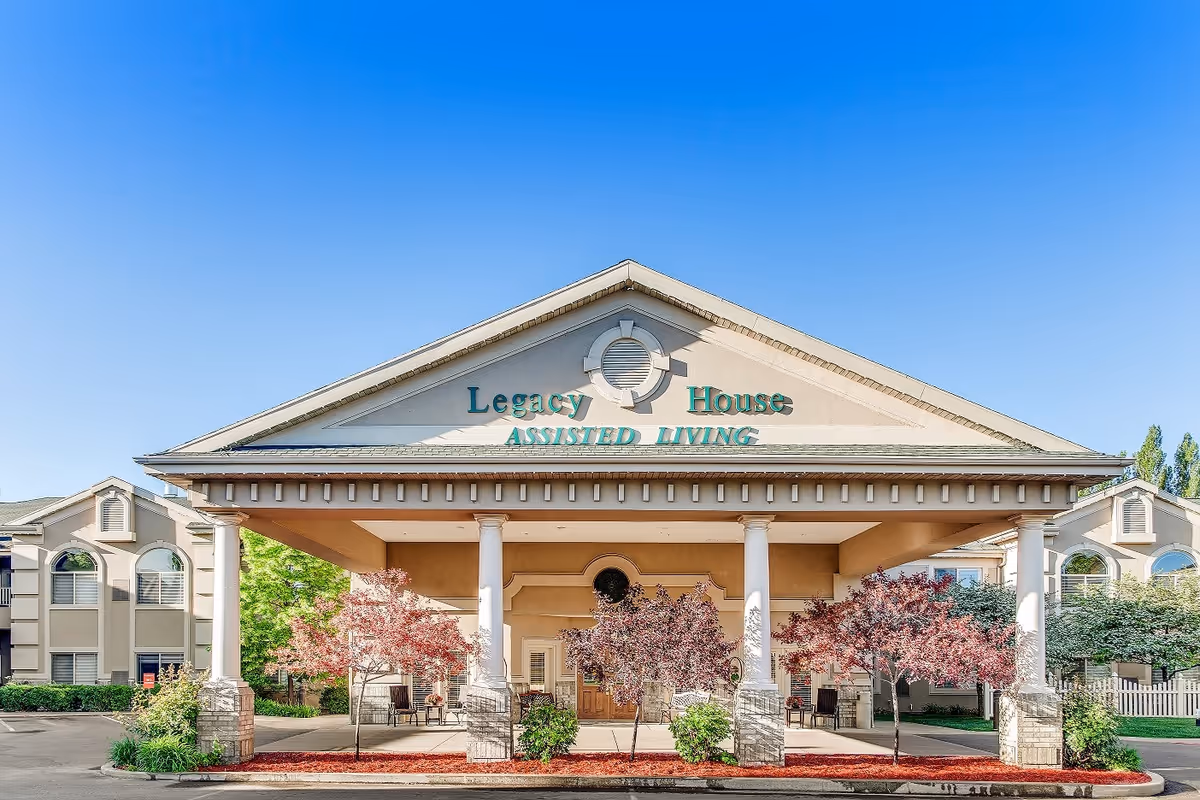 Front exterior view of Legacy House Assisted Living facility with a covered entrance supported by white columns, landscaped with small trees and bushes, under a clear blue sky.