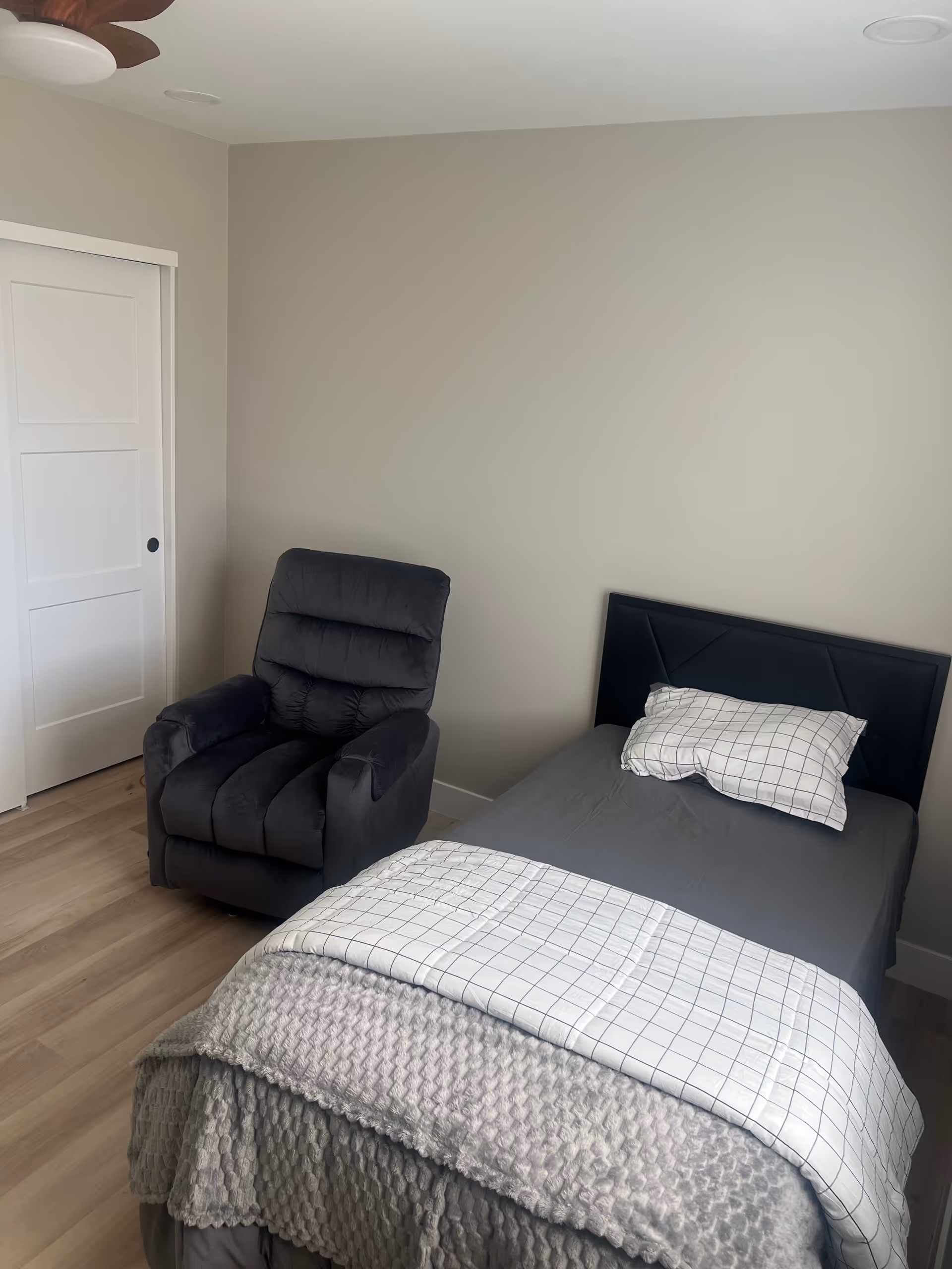 Small bedroom with a single bed dressed in gray and checkered linens and a dark recliner against a beige wall.