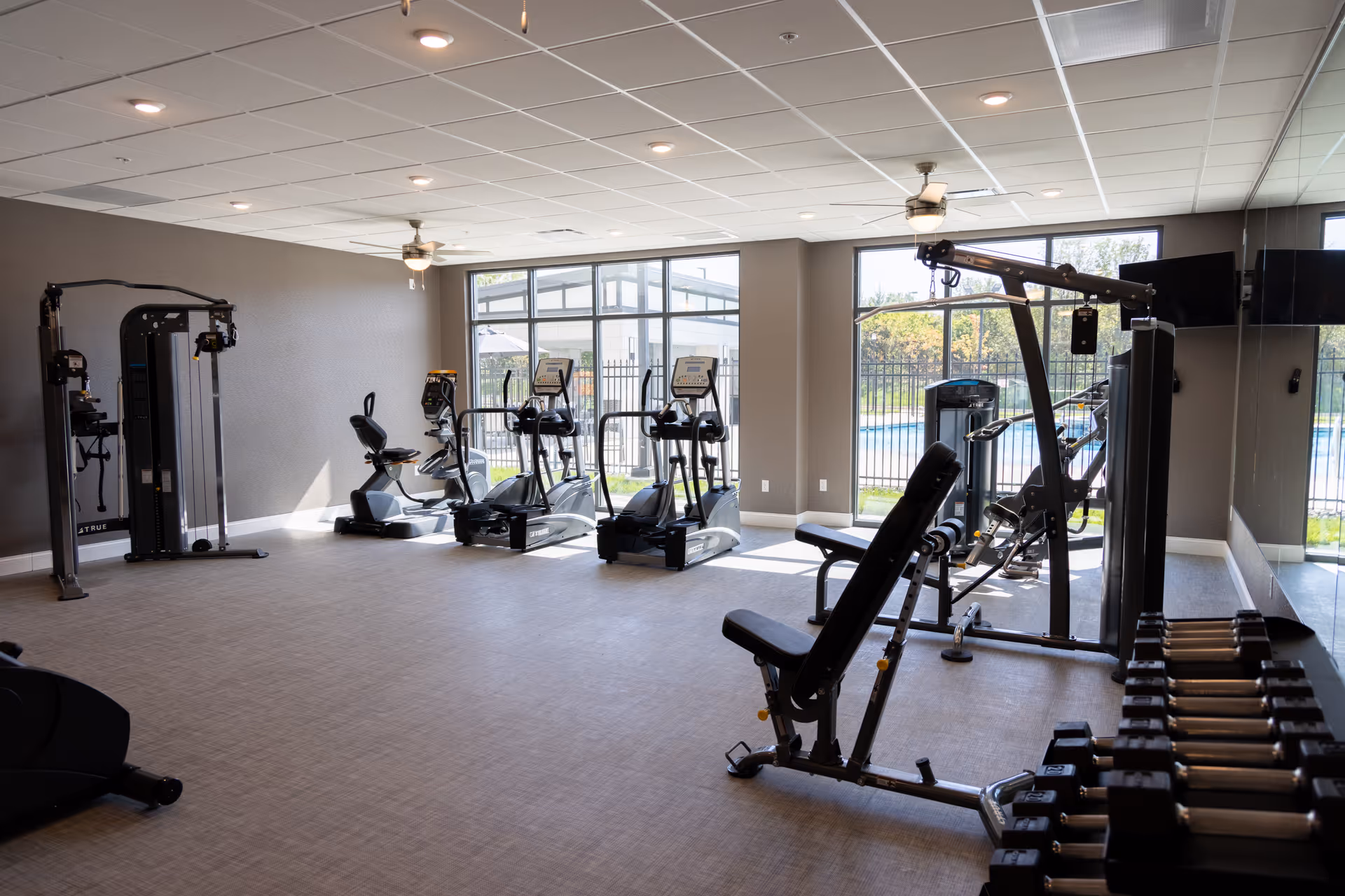 A well-lit fitness room with various exercise equipment including stationary bikes, elliptical machines, weight bench, dumbbells, and a multi-functional weight machine. Large windows provide a view of an outdoor pool area surrounded by a fence.