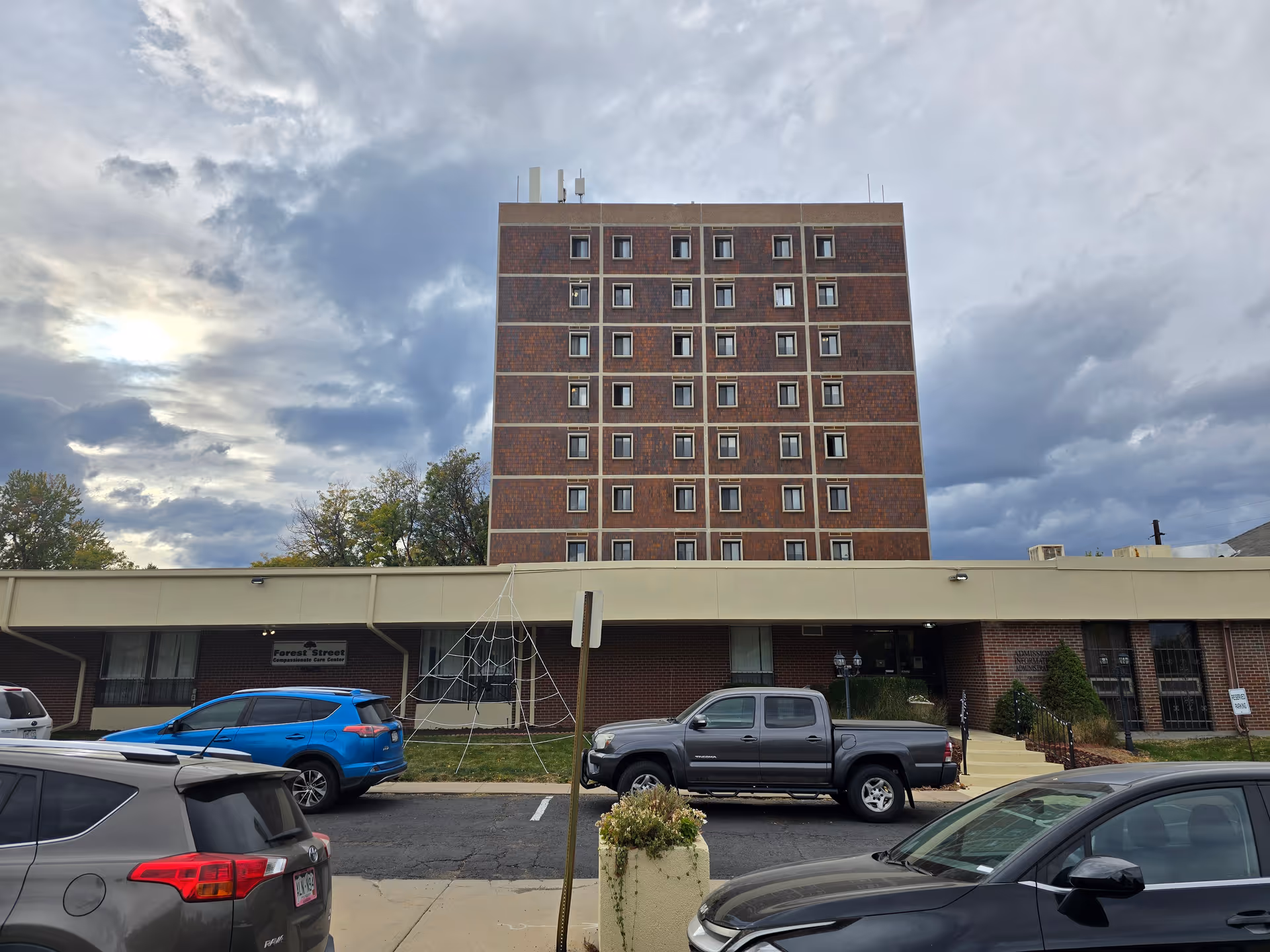 Exterior view of a multi-story brick building with a lower beige section in front, several parked cars, and a cloudy sky overhead. A sign on the building reads 'Forest Street Compassionate Care Center'.