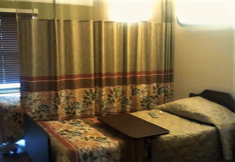 Small nursing facility bedroom with two twin beds separated by a curtain, floral bedspreads, an overbed table and a TV remote.