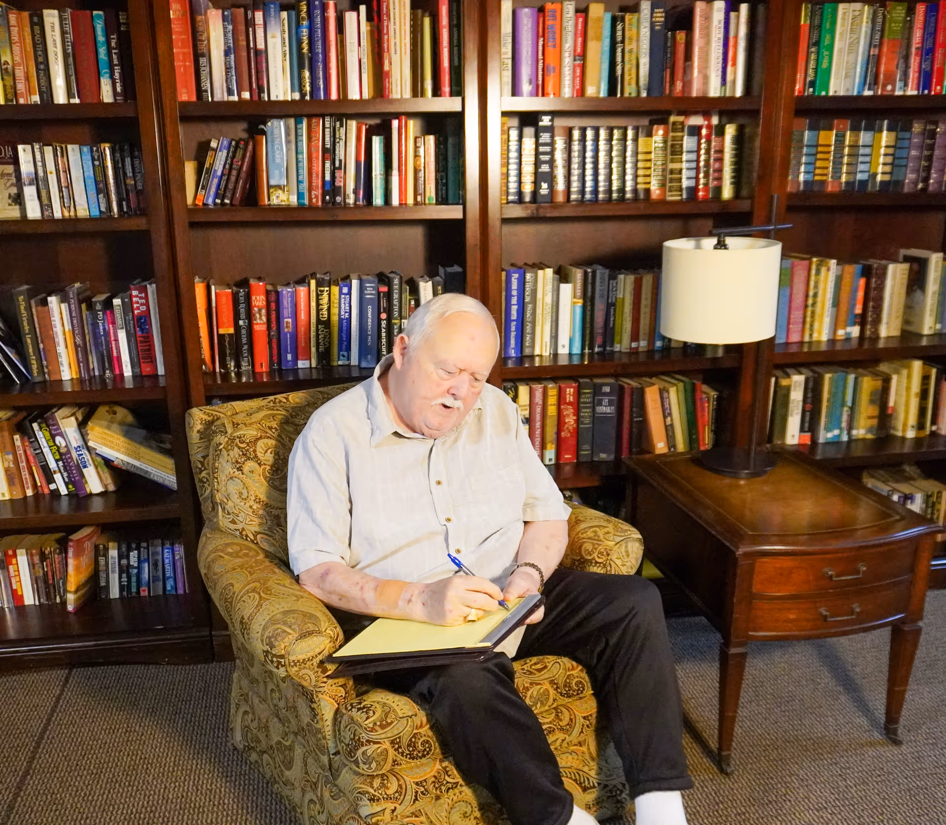 An elderly man sitting in a patterned armchair in a library or reading room, writing on a yellow notepad with a pen. Behind him are wooden bookshelves filled with books, and to his right is a wooden side table with a lamp on it.