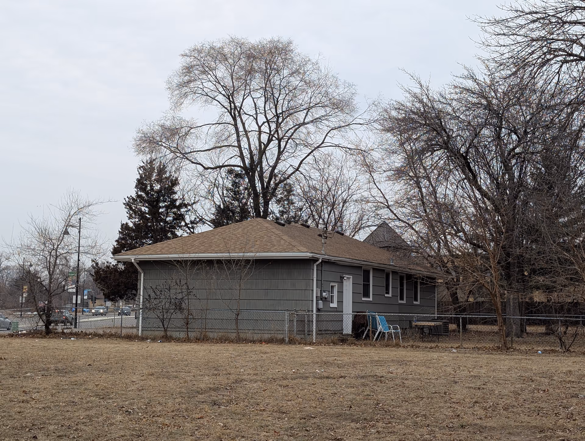 A single-story gray building with a brown shingled roof surrounded by leafless trees and a chain-link fence. There are two blue chairs and a small table near the building. The sky is overcast and the ground is covered with dry grass.