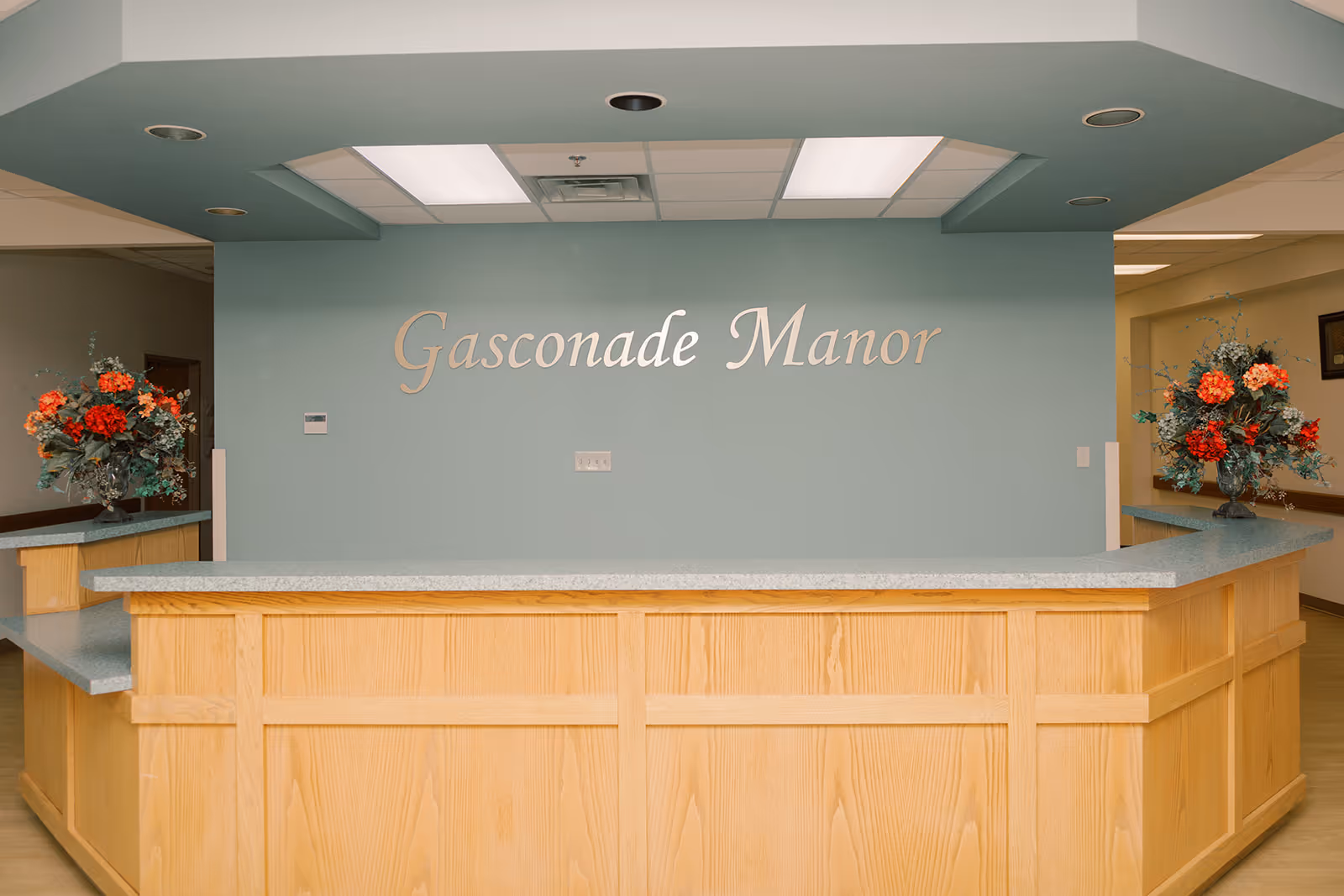 Reception desk area with a teal wall reading "Gasconade Manor" and floral arrangements on either side.