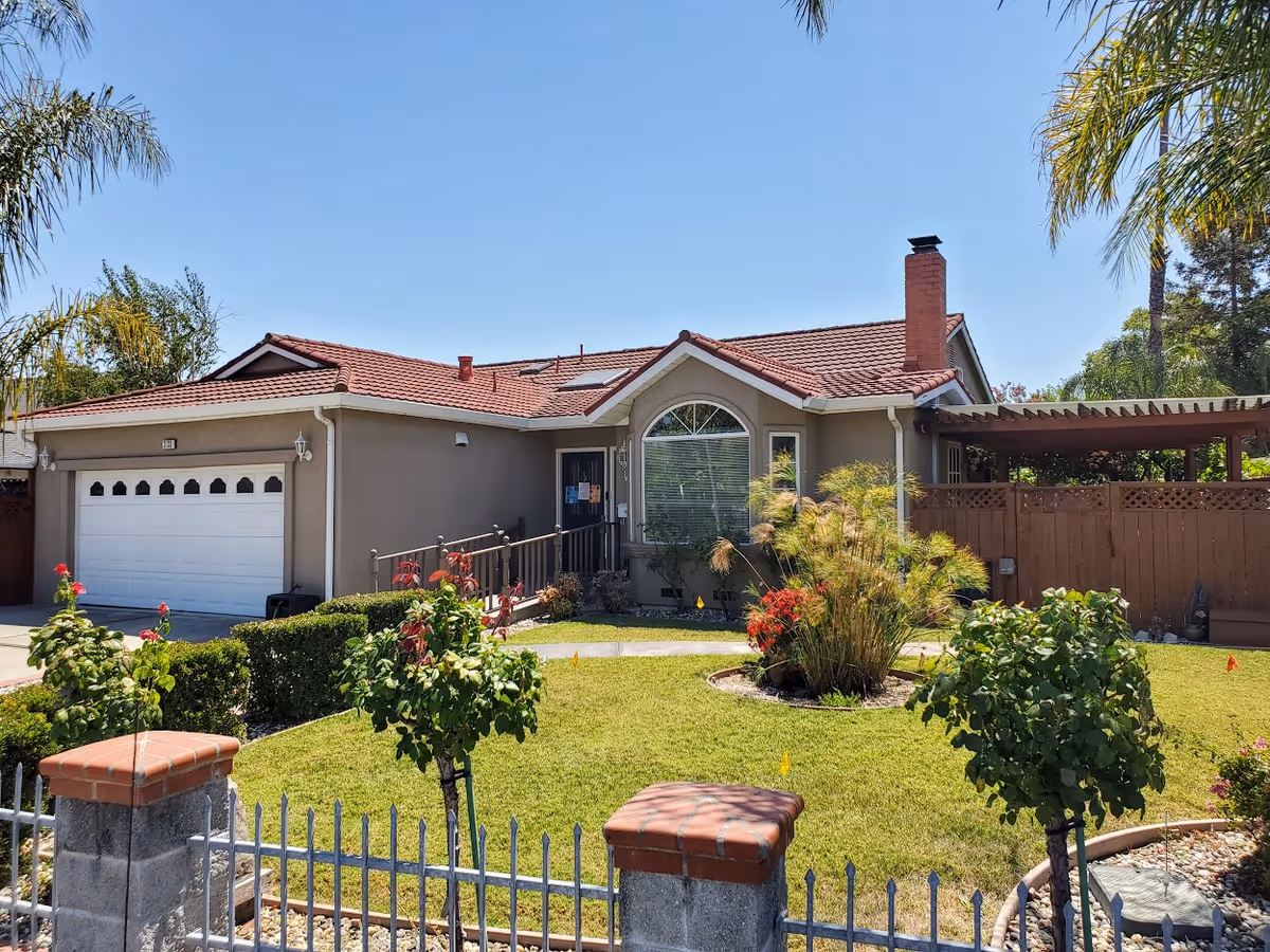 Single-story residential building with a red-tiled roof, a white garage door, and a front yard featuring green grass, bushes, small trees, and a paved walkway leading to the entrance. The yard is enclosed by a metal fence with brick pillars. Palm trees are visible in the background under a clear blue sky.