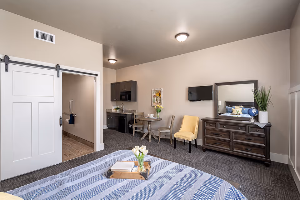 A senior living facility bedroom with a bed in the foreground covered with a striped blue and white blanket and a wooden tray holding a book and a vase of white tulips. The room features a dark wooden dresser with a large mirror, a yellow upholstered chair, a small round table with two chairs, and a kitchenette with dark cabinets, a microwave, and a mini fridge. A sliding barn door on the left leads to a bathroom area.