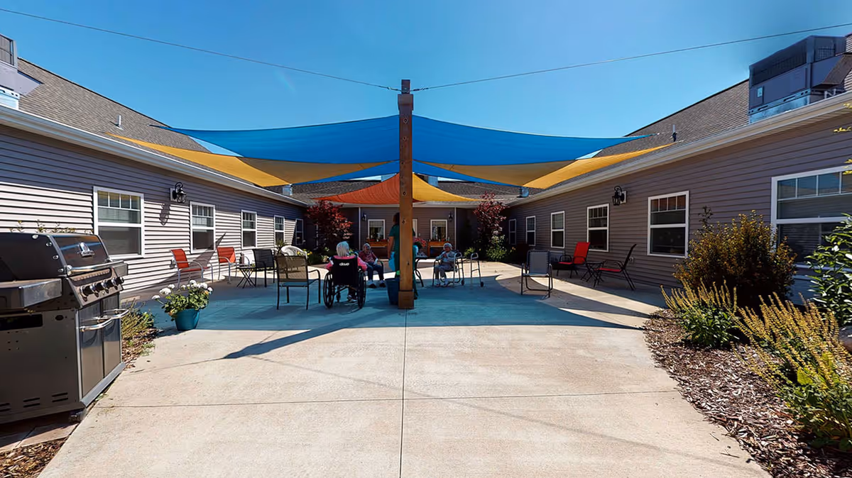 Outdoor courtyard area at Vicinia Gardens Luxury Retirement Living with several seniors sitting on chairs and one in a wheelchair under colorful shade sails. The courtyard is surrounded by single-story buildings with windows and has a barbecue grill on the left side and landscaped plants on the right.