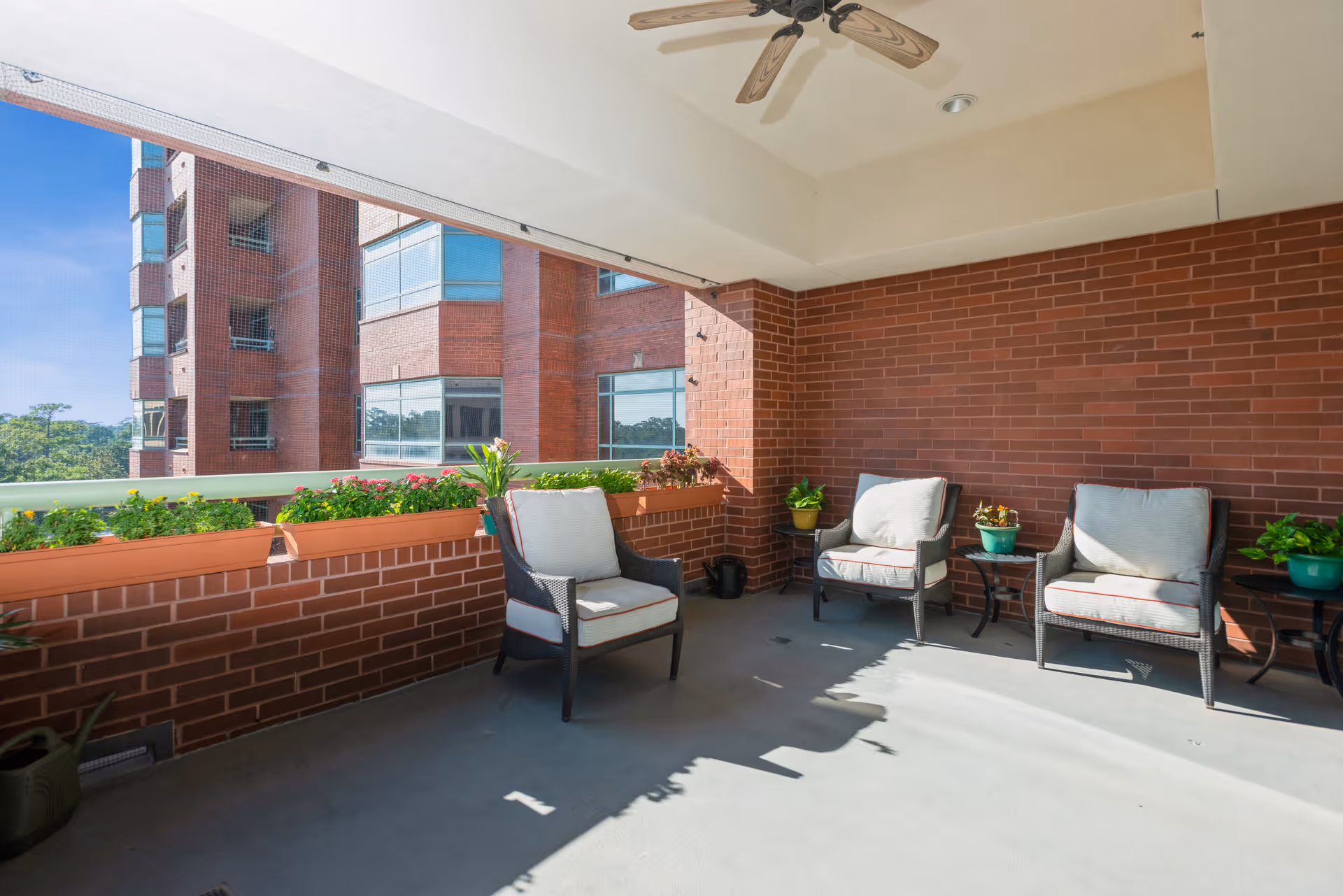 A covered outdoor patio area with three cushioned chairs and small tables with potted plants. The space has brick walls and a ceiling fan, with a view of a nearby brick building and greenery in the background.