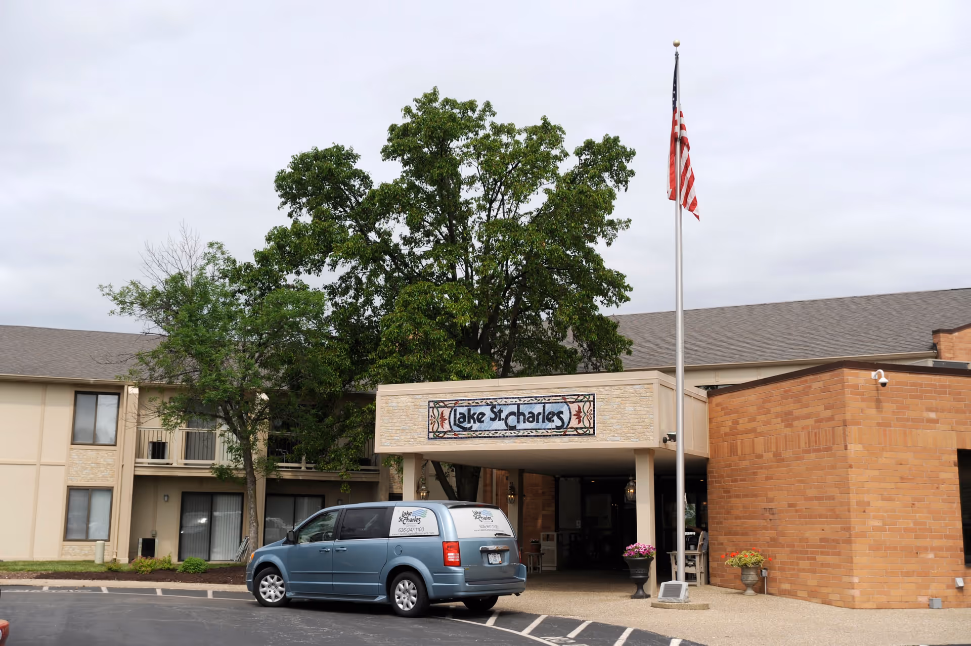 Exterior view of Lake St. Charles Retirement Community building entrance with a covered drop-off area, an American flag on a flagpole, a blue van parked in front with the facility's name on it, and trees around the building.