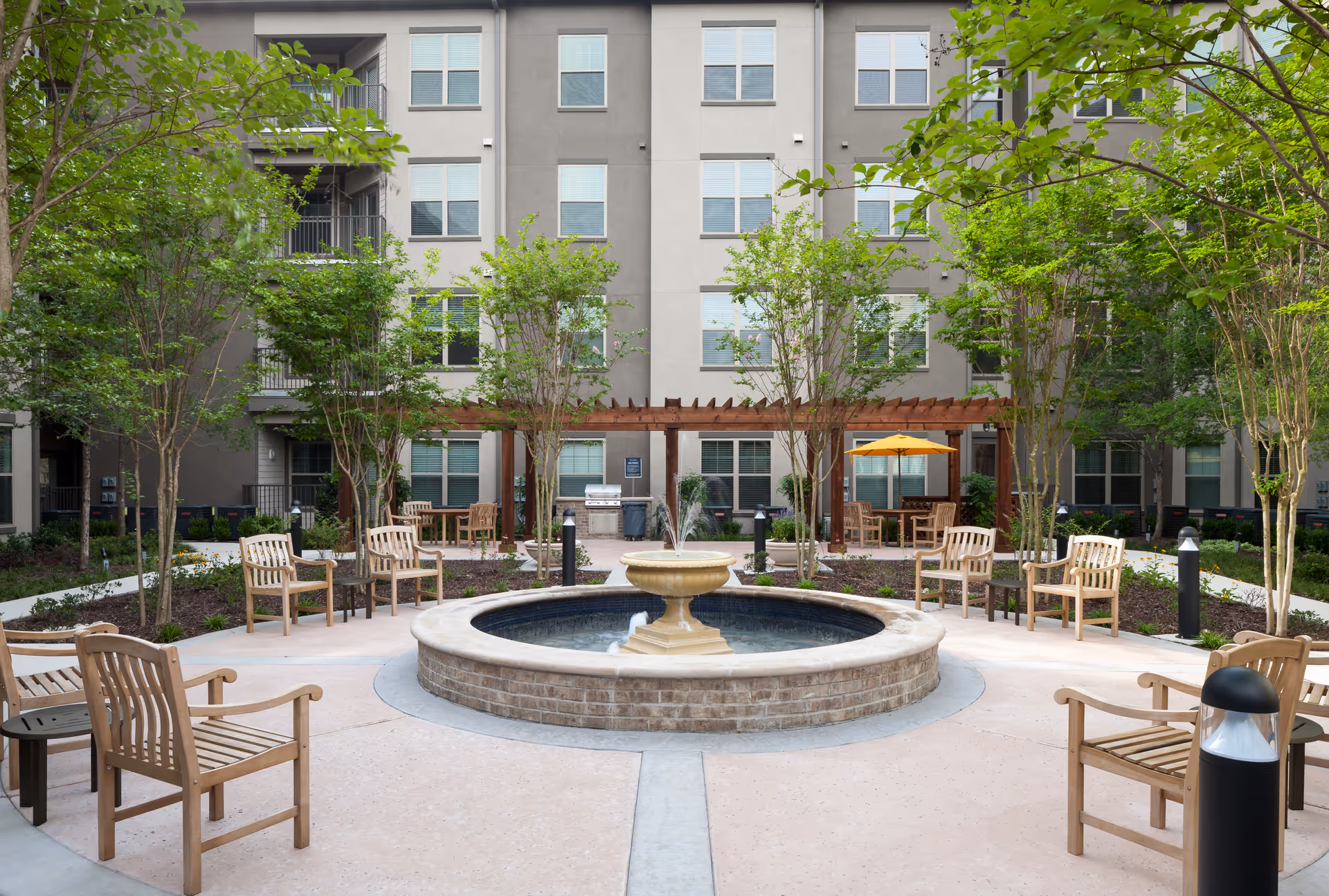 Outdoor courtyard area at Firewheel Town Village featuring a circular stone fountain in the center surrounded by wooden benches and chairs. There are small trees and landscaped garden beds around the seating area, with a pergola and patio tables with umbrellas in the background. The courtyard is enclosed by a multi-story residential building.