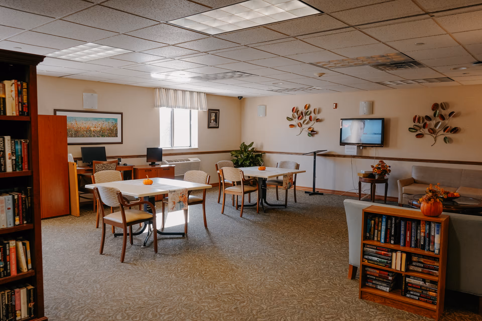 A cozy senior living common area with tables and chairs arranged for socializing or activities. The room features a bookshelf filled with books, a wall-mounted TV, decorative wall art, and a few small pumpkins on the tables and shelves, suggesting a fall or autumn theme. There is a window letting in natural light and a computer desk in the corner.