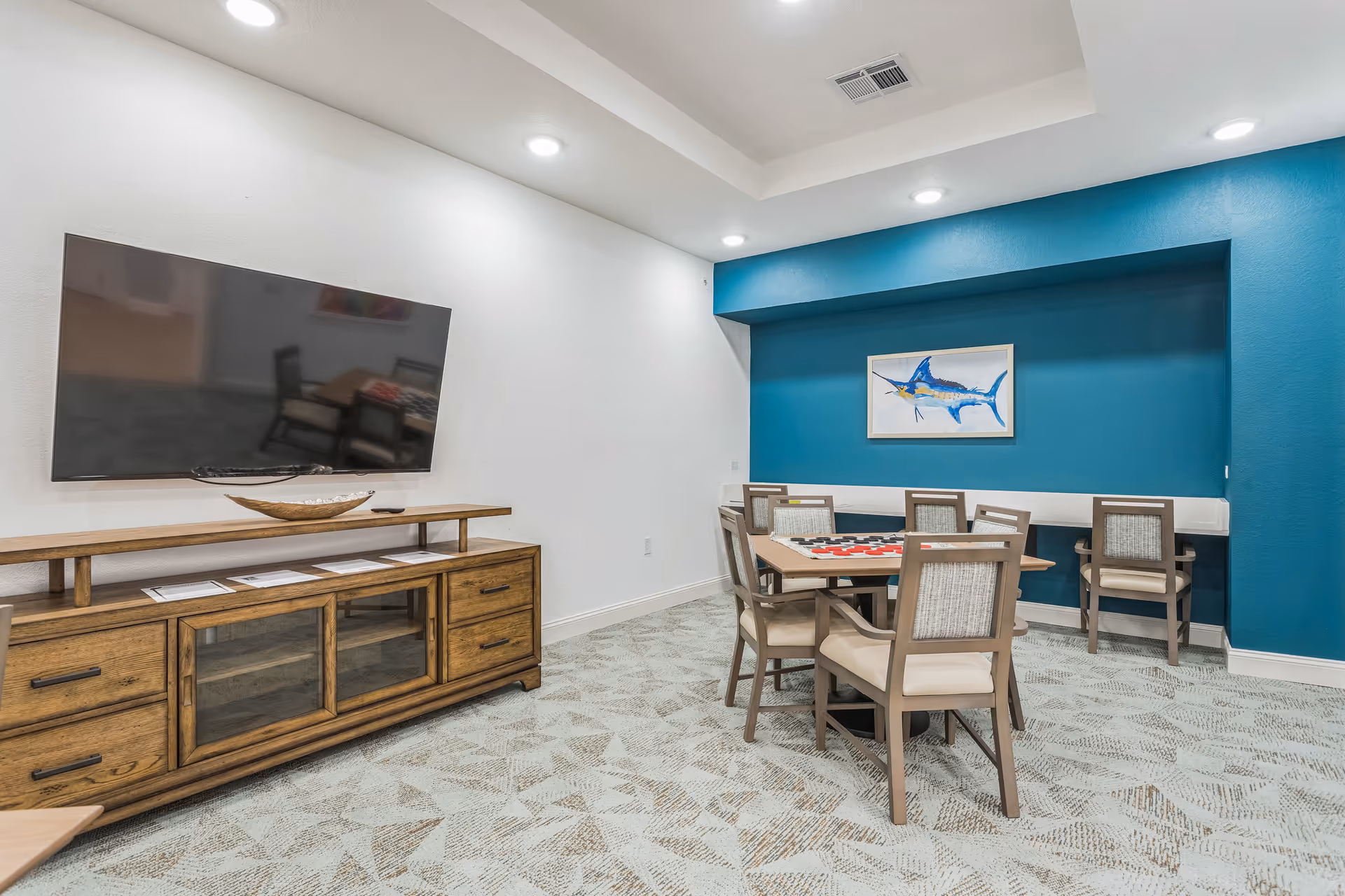 A bright communal living room with a wall-mounted TV, wooden media console, and tables with chairs set against a teal accent wall.
