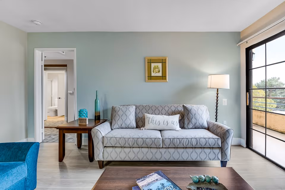 A cozy living room with a patterned gray sofa adorned with three cushions, one of which says 'this is us'. To the left of the sofa is a wooden side table with decorative items including a tall green bottle and a small blue vase. A floor lamp with a white shade stands to the right of the sofa. There is a large sliding glass door on the right side leading to a balcony with a view of trees. In the background, an open doorway reveals a glimpse of a bathroom. The walls are painted light green and the floor is light wood.