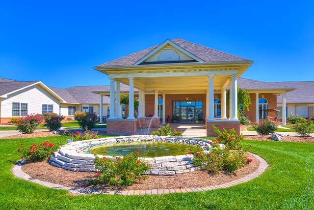 Front exterior view of Cedarhurst Senior Living of Granite City featuring a large covered entrance with white columns, a circular stone fountain surrounded by flowers and greenery, and a clear blue sky overhead.