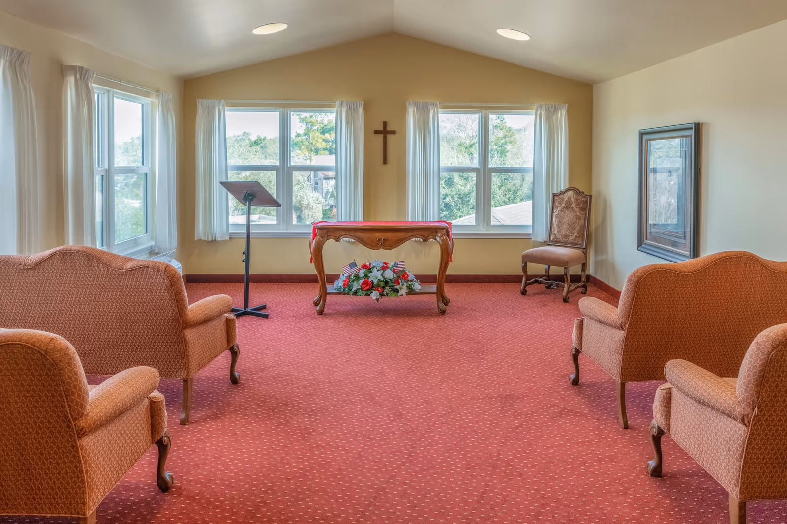 A small chapel or meditation room with four upholstered chairs arranged facing a wooden table with a floral arrangement underneath. The room has large windows with white curtains, a cross mounted on the wall, a music stand, and a framed picture on the right wall. The carpet is red with a subtle pattern.