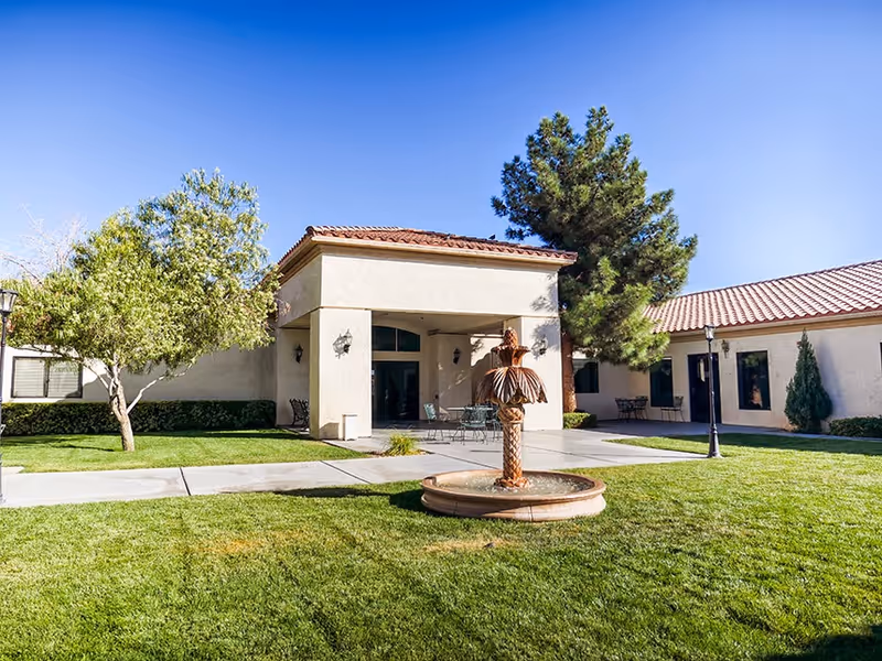 Exterior view of Silver Ridge Healthcare Center showing a beige building with a tiled roof, a covered entrance area with outdoor seating, a decorative fountain in the foreground, green grass, trees, and a clear blue sky.