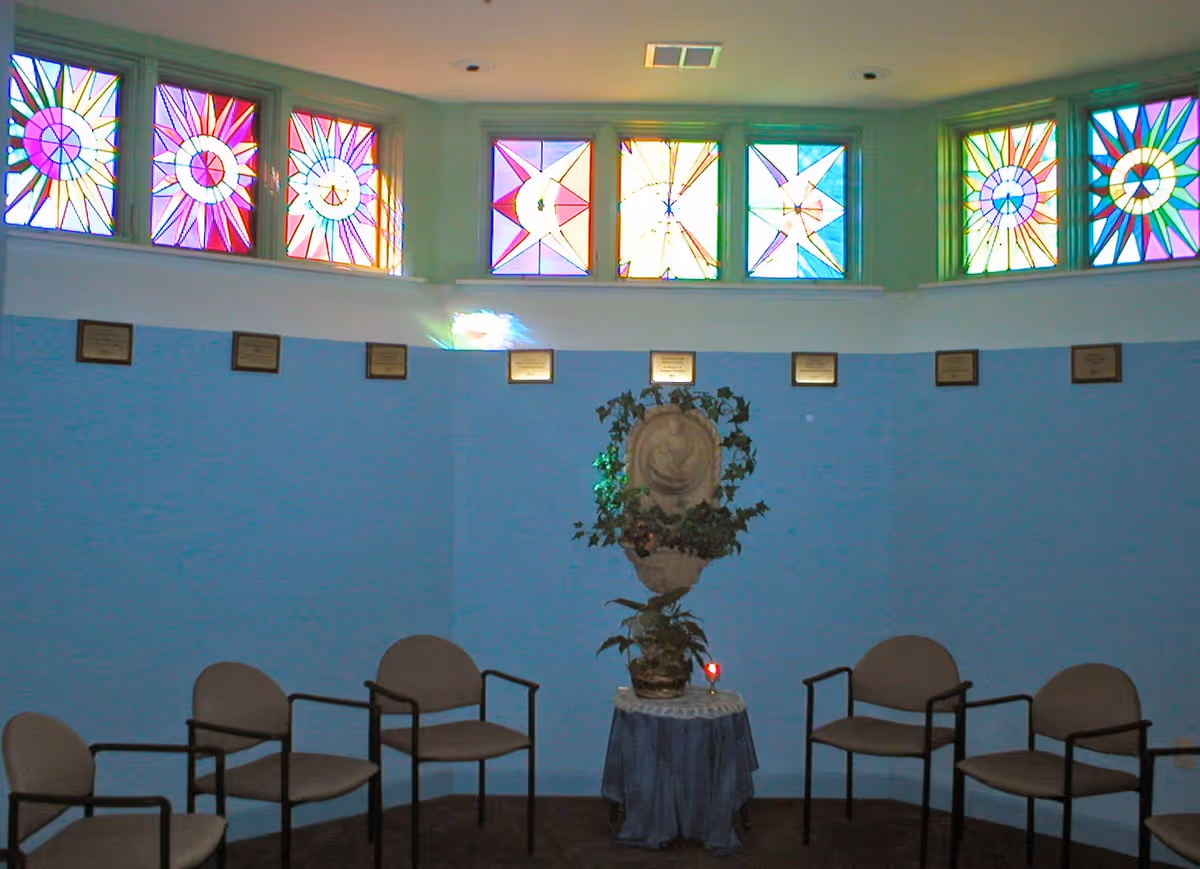 A small circular room with light blue walls and a row of colorful stained glass windows near the ceiling. The room contains six beige chairs arranged in a semicircle around a small table covered with a blue cloth. On the table is a decorative stone sculpture with green plants around it.