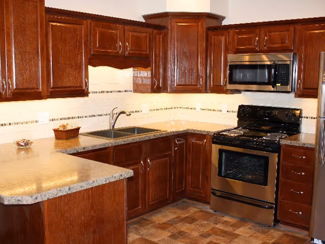 A kitchen with dark wood cabinets, a double sink, a stainless steel oven with a black stovetop, a stainless steel microwave above the oven, and a light-colored countertop with a small basket and a bowl on it. The backsplash is white with a decorative tile strip, and the floor has a patterned tile design.