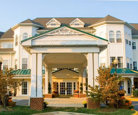 Front exterior view of a large, elegant senior living facility building with a covered entrance supported by white columns and brick bases, surrounded by small trees and landscaping under a clear sky.