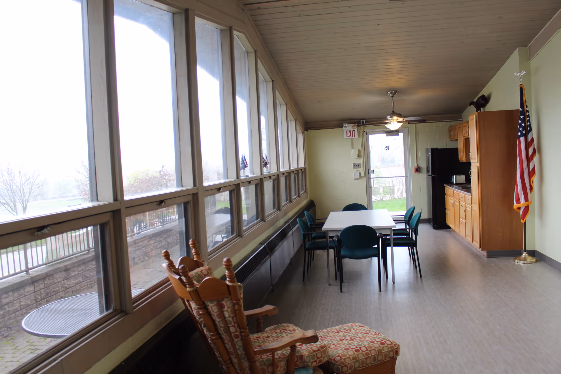 A bright room with large windows along one side, featuring a wooden rocking chair with patterned cushions in the foreground. In the middle of the room is a white table surrounded by six green chairs. At the far end, there is a kitchenette with wooden cabinets, a black refrigerator, a toaster, and an American flag standing in the corner. An exit door with a glass window is visible at the back.
