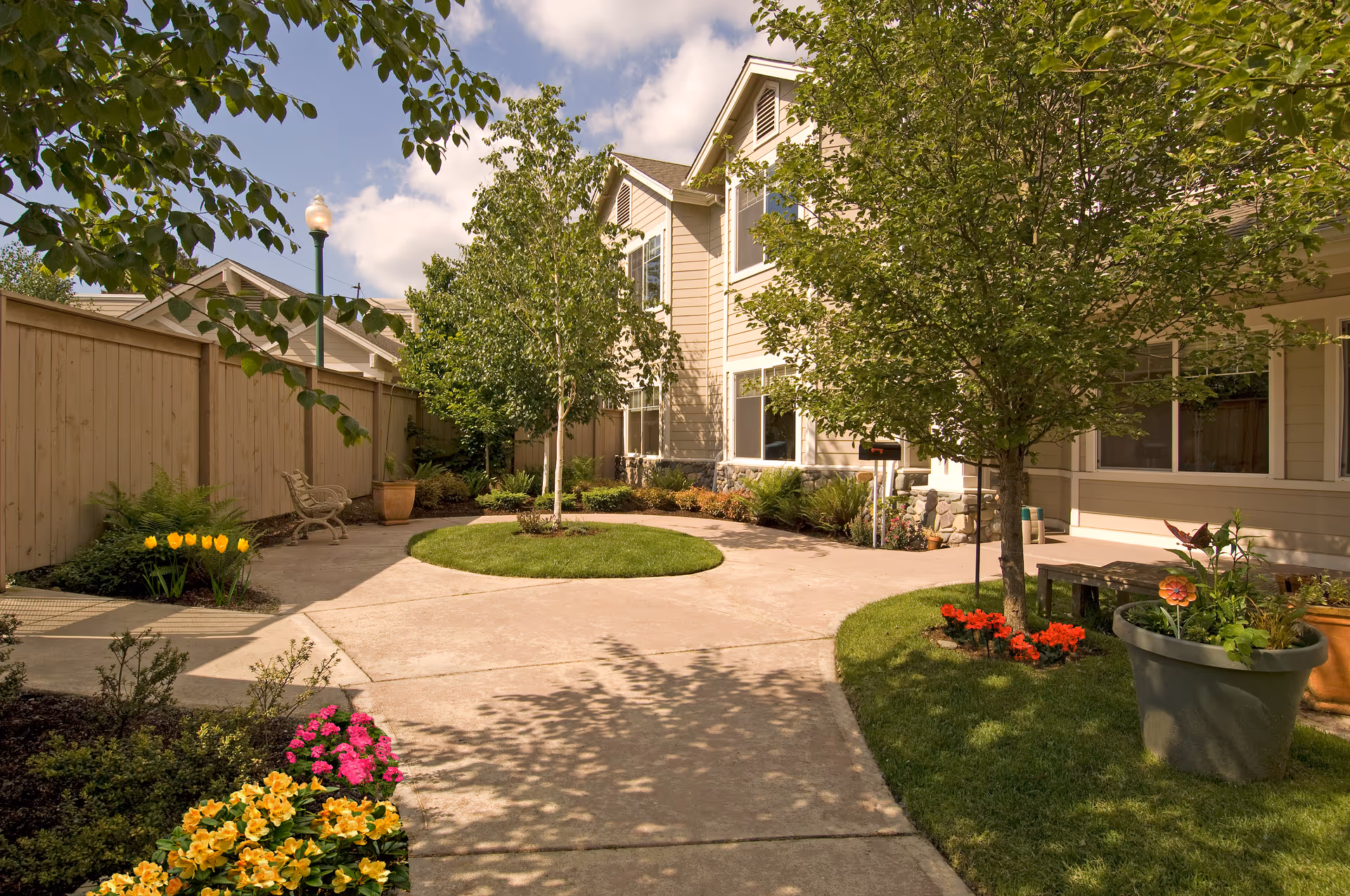 A sunny outdoor courtyard area with a concrete pathway surrounded by green grass, colorful flower beds, and several trees. There is a beige two-story building with multiple windows in the background and a wooden fence on the left side. A bench and a street lamp are also visible along the pathway.