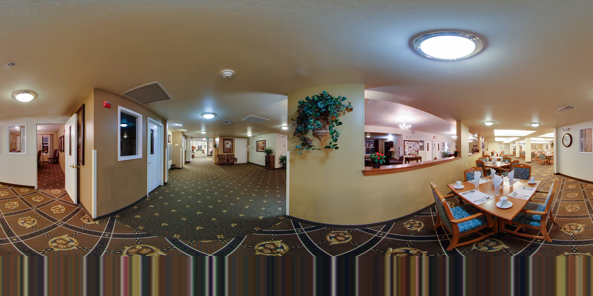 Wide panoramic view of an interior hallway and dining area in a senior living facility. The hallway has beige walls, patterned carpet, and several doors leading to rooms. The dining area features wooden tables set with white napkins, cups, and plates, surrounded by blue upholstered chairs. There are plants and framed pictures on the walls, and ceiling lights illuminate the space.