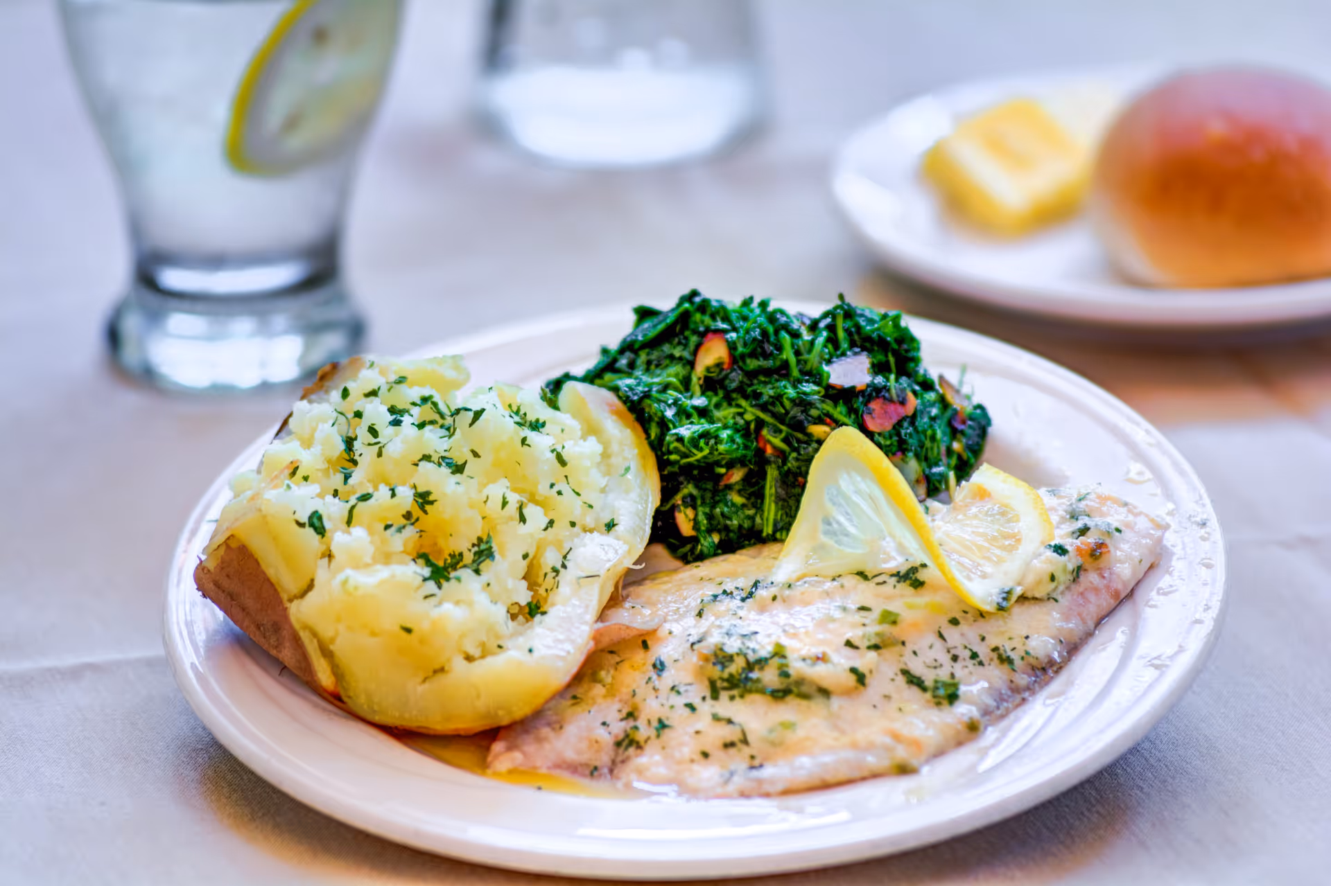 A plate of food featuring a baked potato topped with herbs, sautéed spinach with garlic slices, and a fillet of fish garnished with lemon slices. In the background, there is a glass of water with a lemon slice and a plate with a bread roll and butter.