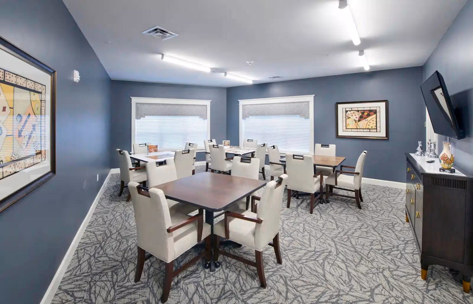 A dining room with several square wooden tables and beige upholstered chairs with wooden armrests. The walls are painted blue and decorated with framed abstract artwork. Two large windows with white blinds and valances provide natural light. A dark wooden sideboard with decorative items and a wall-mounted flat-screen TV are on the right side of the room. The floor is carpeted with a patterned design.