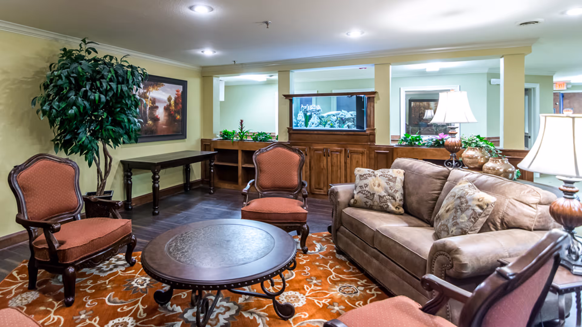 A cozy common lounge with a sofa, upholstered armchairs, a round coffee table, lamps, decorative rug, and an aquarium.