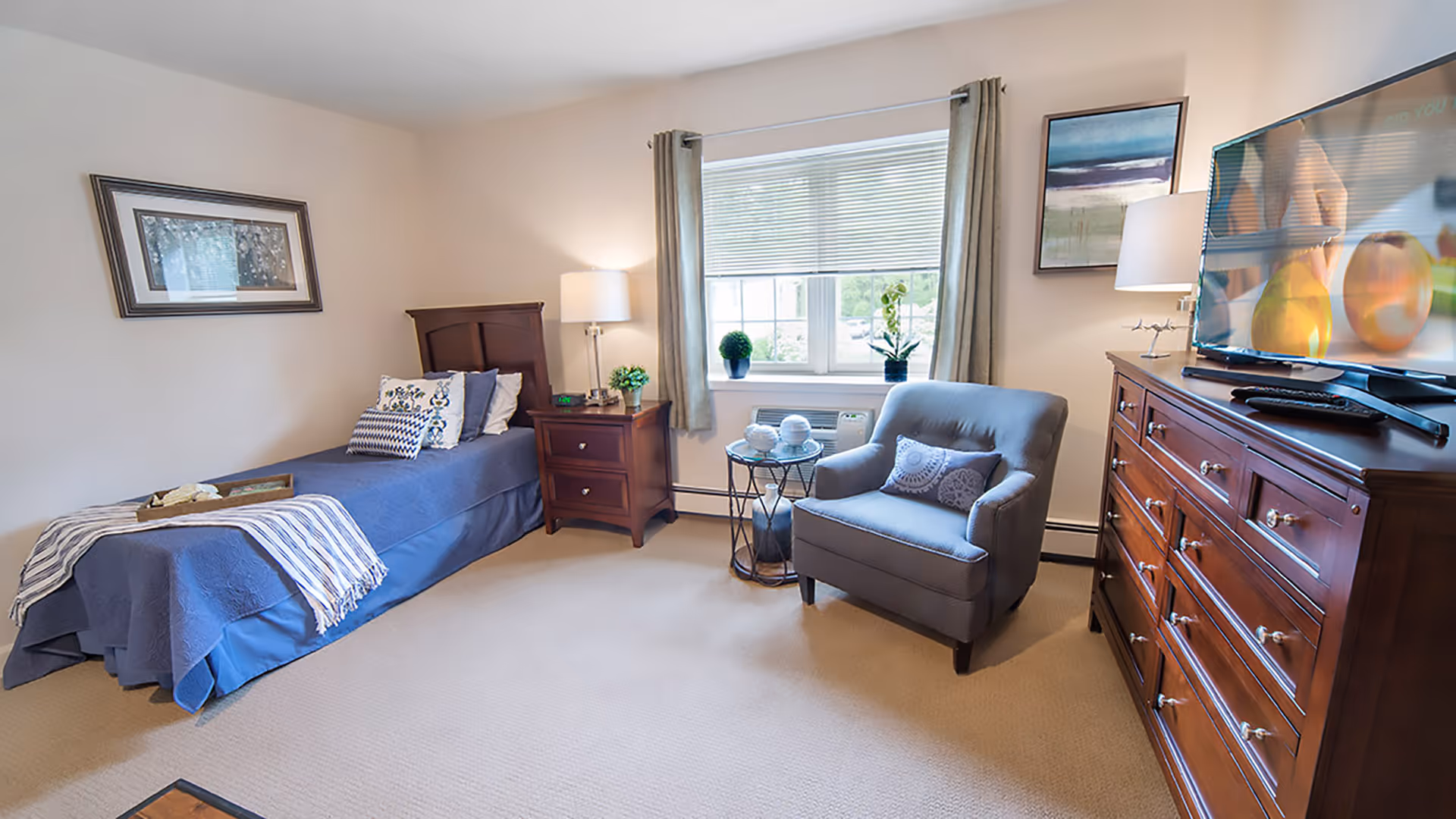 A cozy senior living bedroom featuring a single bed with blue bedding and decorative pillows, a wooden nightstand with a lamp and small plant, a comfortable gray armchair with a patterned pillow, a round side table with decorative items, a large wooden dresser with a flat-screen TV on top, and a window with beige curtains letting in natural light.