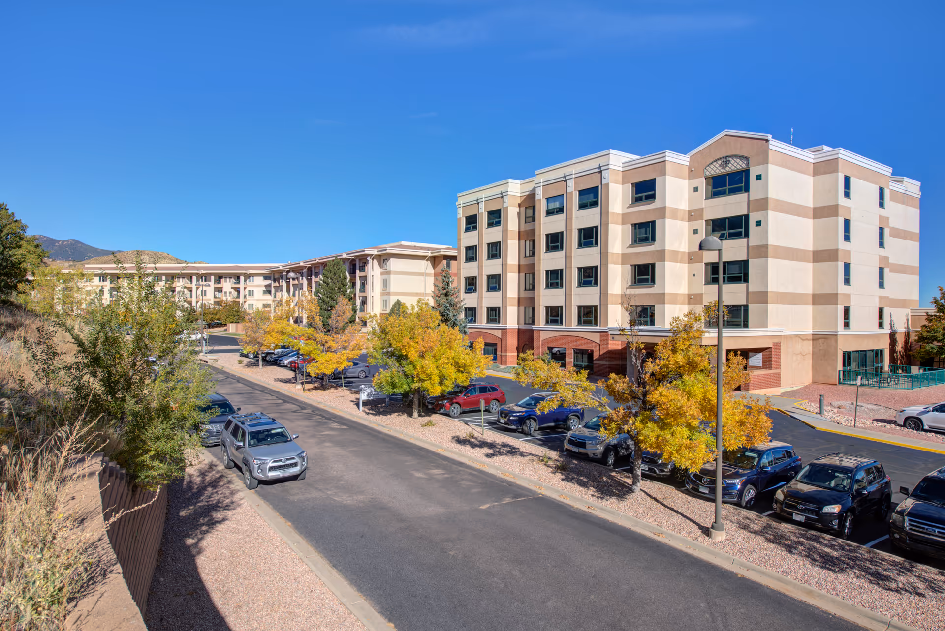 Exterior view of a multi-story senior living facility named Brookdale Skyline with a parking lot in front, several parked cars, trees with autumn foliage, and a clear blue sky.