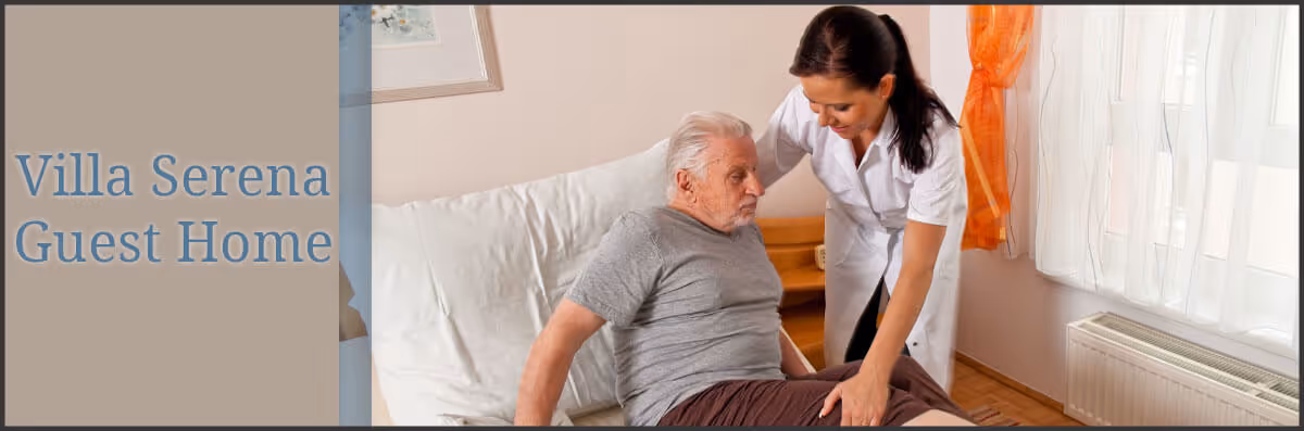 A caregiver assisting an elderly man sitting on a bed in a softly lit room with a window covered by sheer white curtains and orange drapes.