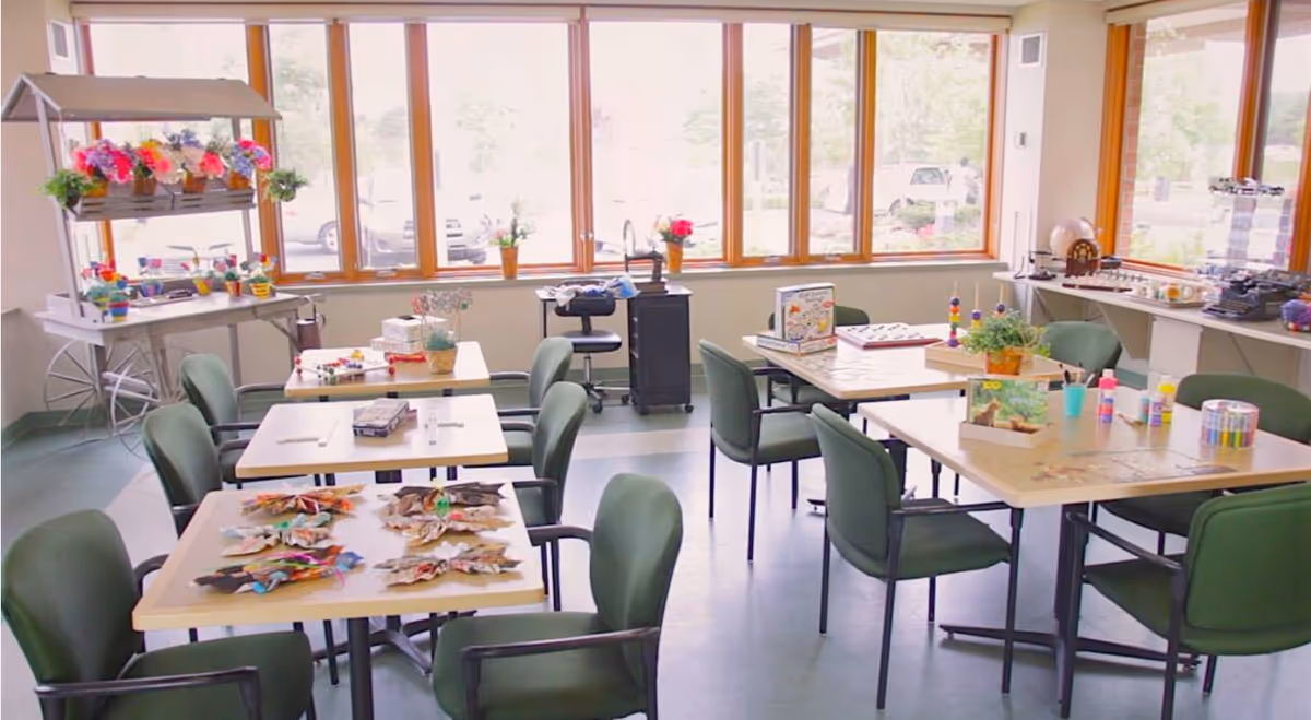 A bright activity room with several tables and green chairs arranged around them. The tables have various craft supplies, games, and decorations on them. Large windows let in natural light, and there is a cart with colorful potted flowers near the windows.