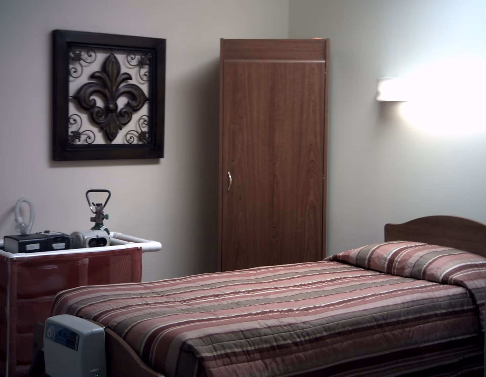 A simple bedroom with a single bed covered in a striped bedspread in shades of brown and beige. There is a wooden wardrobe against the wall, a decorative wall hanging with a fleur-de-lis design, and medical equipment on a small table next to the bed. A wall-mounted light fixture illuminates the room.