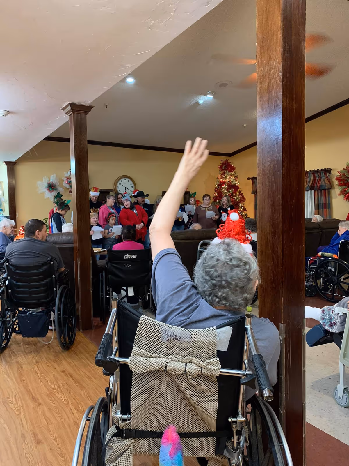 A group of elderly people, many in wheelchairs, gathered in a common room decorated for Christmas with a Christmas tree and wreaths. Some people are standing and singing, while one person in the foreground is raising their hand. The room has wooden pillars and a clock on the wall.