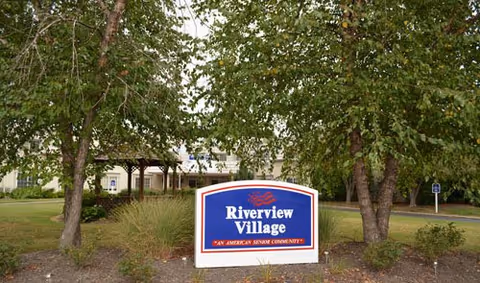 Entrance sign reading "Riverview Village" in front of trees with the facility building visible behind.