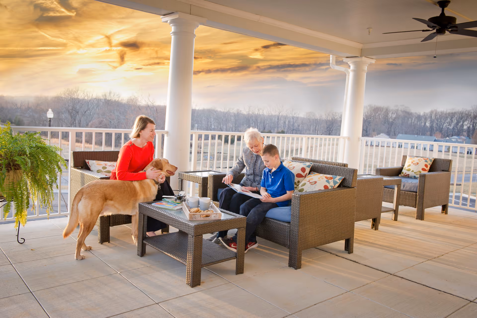 A covered outdoor patio area at sunset with wicker furniture including a sofa and chairs with floral cushions. An elderly woman and a young boy are sitting on the sofa looking at a book, while a woman in a red sweater pets a golden retriever dog standing nearby. There is a potted fern plant on the left side and a scenic view of trees and houses in the background.