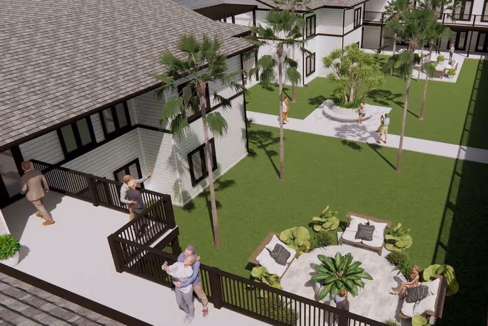 Aerial view of an outdoor courtyard area at a senior living facility with green lawns, palm trees, and seating areas with cushioned chairs around a circular paved space. Several people are walking or standing on the pathways and balconies of the white buildings with dark trim surrounding the courtyard.