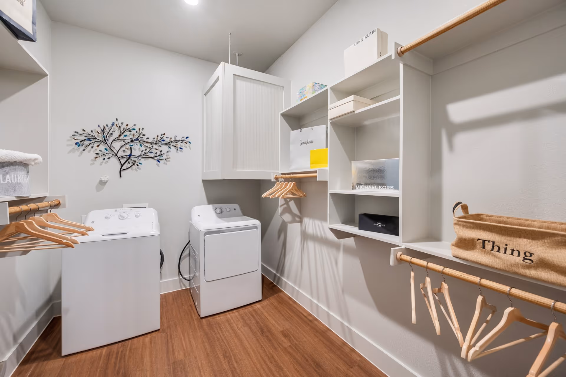 Bright laundry room with a washer and dryer, open shelving, hanging rods with wooden hangers, and decorative wall art.