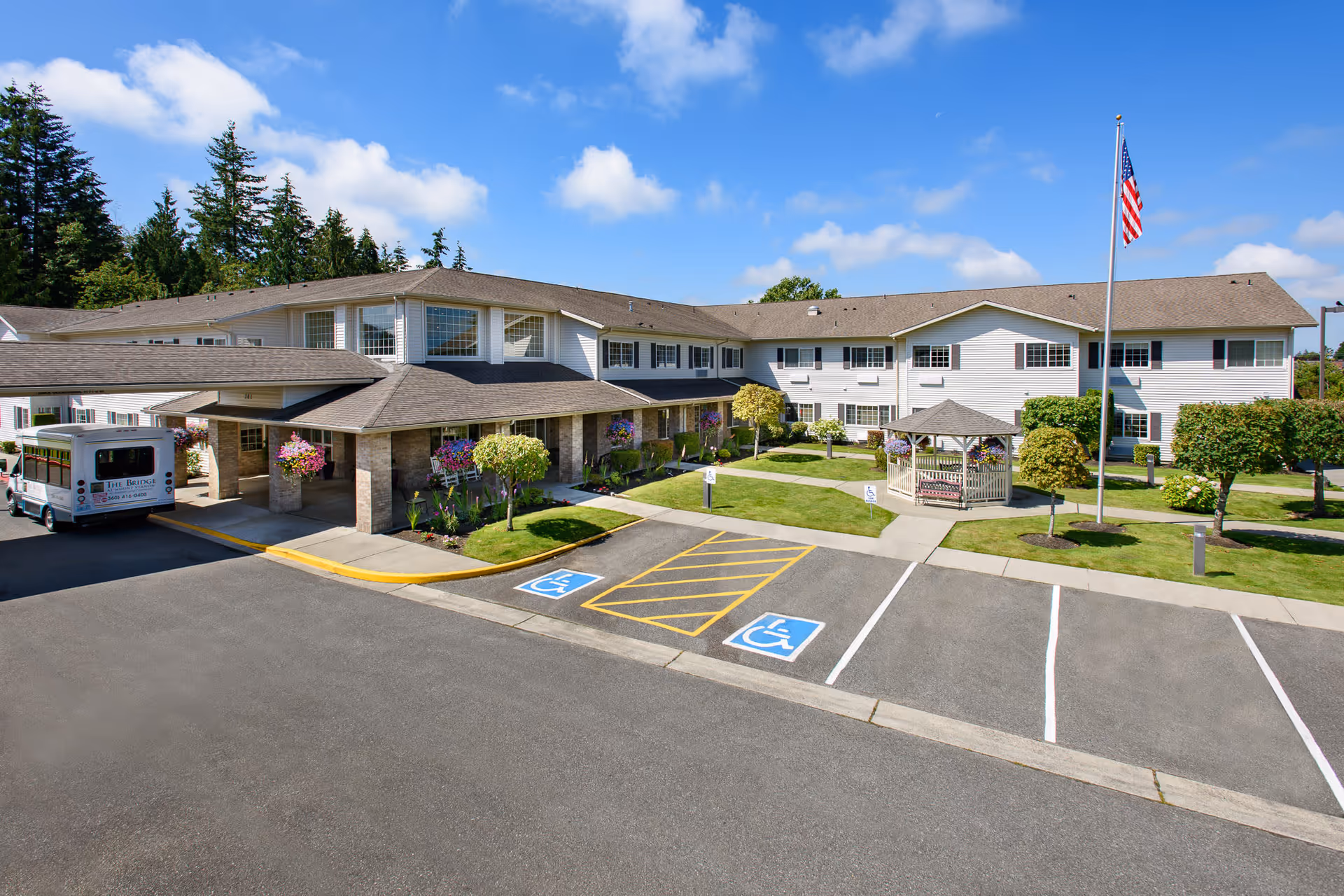 Exterior view of The Bridge at Mount Vernon senior living facility on a sunny day, showing a two-story building with white siding and multiple windows, a covered entrance with hanging flower baskets, a small gazebo in a landscaped courtyard, an American flag on a flagpole, and a parking lot with handicap spaces.